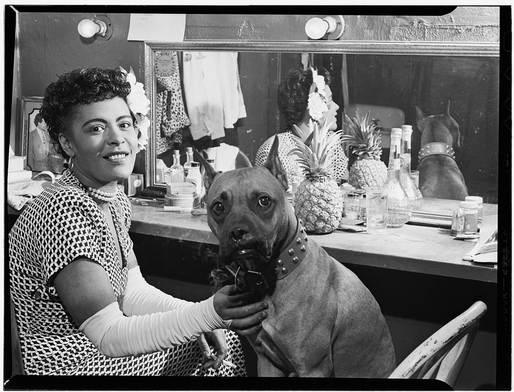 Retrato de mulher negra com luvas brancas senta em frente ao espelho ao lado do seu cachorro de estimação, ambos olhando para a câmera