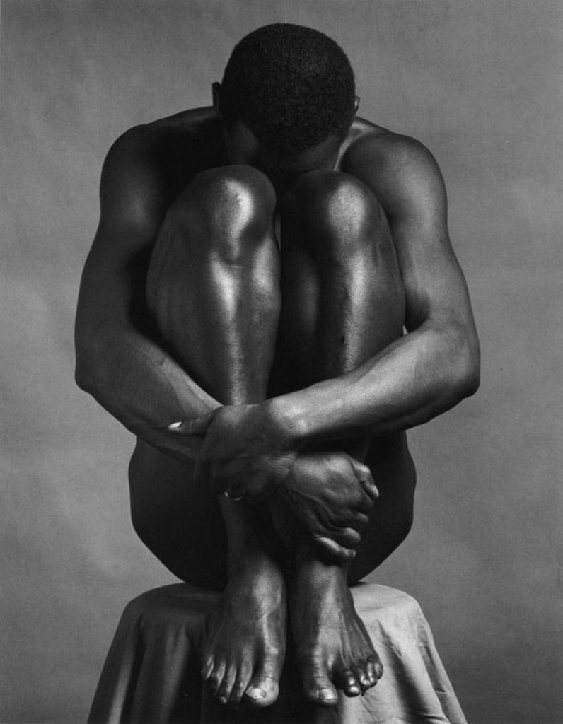 "Ajitto" (1981), do fotógrafo Robert Mapplethorpe. Homem negro nu encolhido com os braços envolvendo suas pernas, num banco coberto por tecido. Imagem em preto e branco.