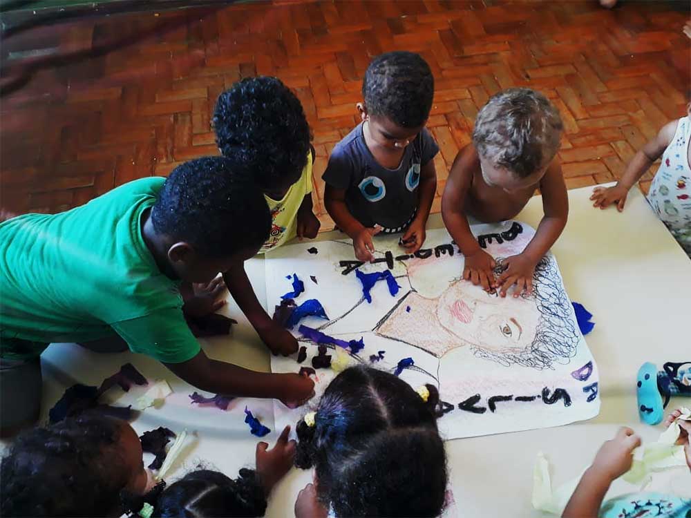 Foto colorida mostra crianças fazendo uma atividade escolar de colagem da Benedita da Silva no Colegio Maria Gregoria
