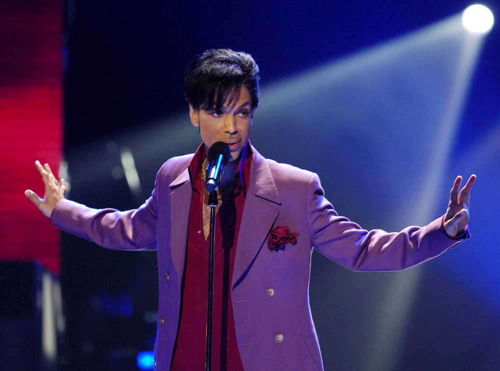 Prince no palco, posando com braços abertos, veste paletó roxo sobre camisa de cor bordô escuro