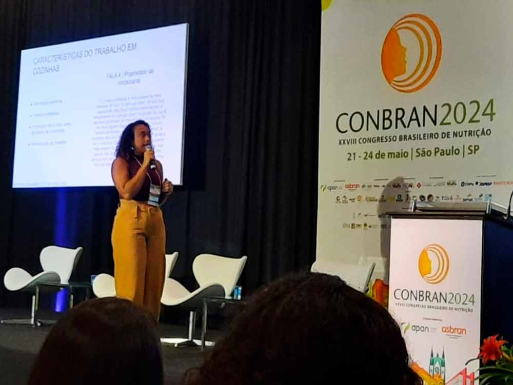 Rayane de Freitas palestra no palco, com projeção ao fundo e grande painel da CONBRAN ao lado.