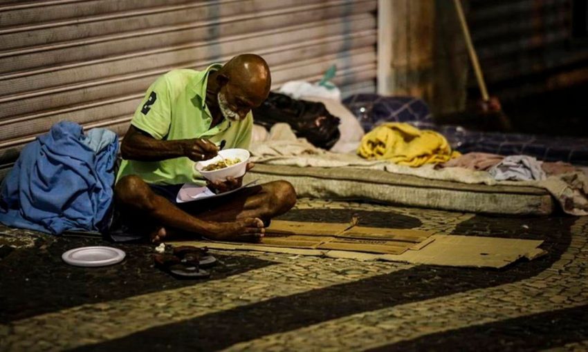 Morador de rua negro jantando em meio a seus pertences na calçada. (Foto: Agência O Globo)